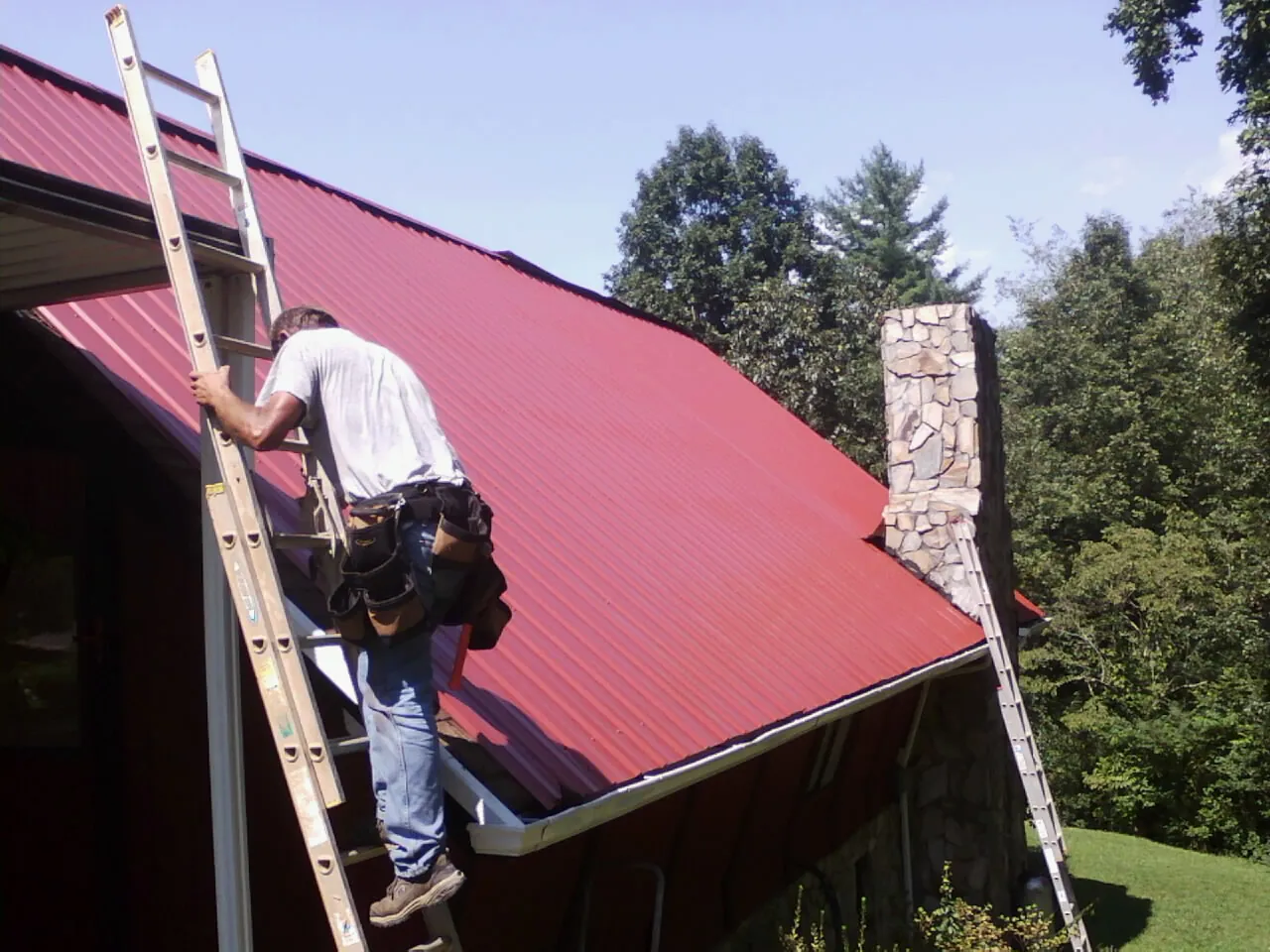Roofer on a ladder beside a red metal roof and stone chimney — Soffit & Fascia Repair by Roofing Brothers in Emerado